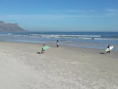 a group of people walking on the beach with surfboards at Beach Haven Sea Views, Fritzandi, Strand, Cape Town in Cape Town