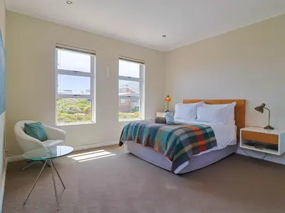 a bedroom with a bed and a chair and two windows at Penguin View in Bettyʼs Bay