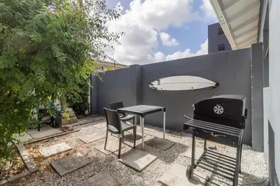 a grill and a table and a surfboard against a fence at 1Bed Flat, Braai, AC, Walking Distance from Blouberg Beachfront in Cape Town
