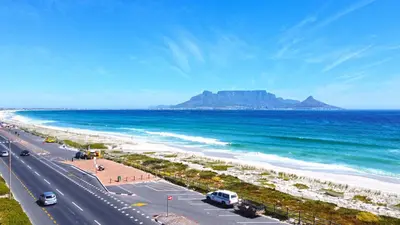 a road next to the beach with a mountain in the background at SeeHUYS Boutique Bloubergstrand Beachfront Studio in Bloubergstrand