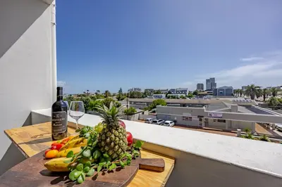 a plate of fruit on a table on a balcony with a bottle of wine at Contemporary Modern 1 Bedroom-19B, Blouberg in Cape Town