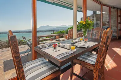 a wooden table and chairs on a balcony with a view of the ocean at 18 On Kloof Guest House in Gordonʼs Bay
