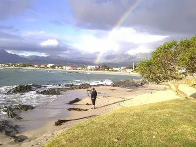 a man fishing on the beach with a rainbow in the background at 'Die Braaihuis'- sleeps 6, Large Veranda, Sea Views, Central and Fabulous! in Gordonʼs Bay