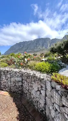 a stone wall with flowers and mountains in the background at Kallistes Misty Shores Cottage in Bettyʼs Bay