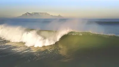 a wave in the ocean with a mountain in the background at Surfhostel Cape Town in Cape Town